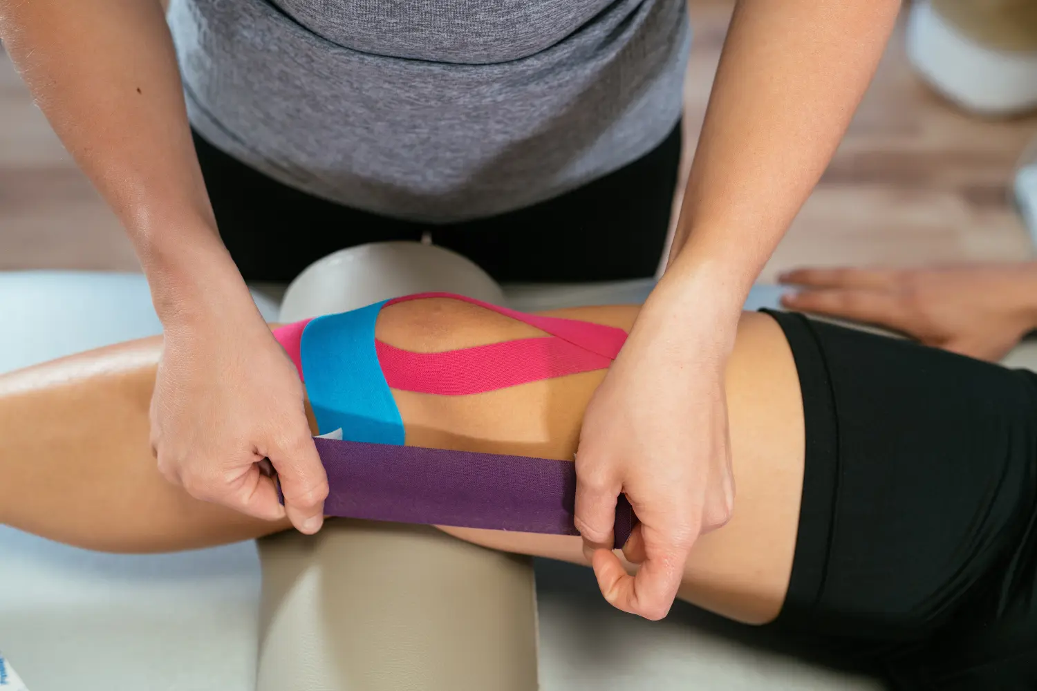 Eine Physiotherapeutin klebt ein lila medizinisches Tape auf ein anderes himmlisches Tape und ein weiteres rosa Tape auf das Knie eines Patienten